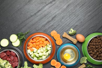 Photo of Natural and dry pet food in feeding bowls among products on dark textured table, flat lay. Space for text