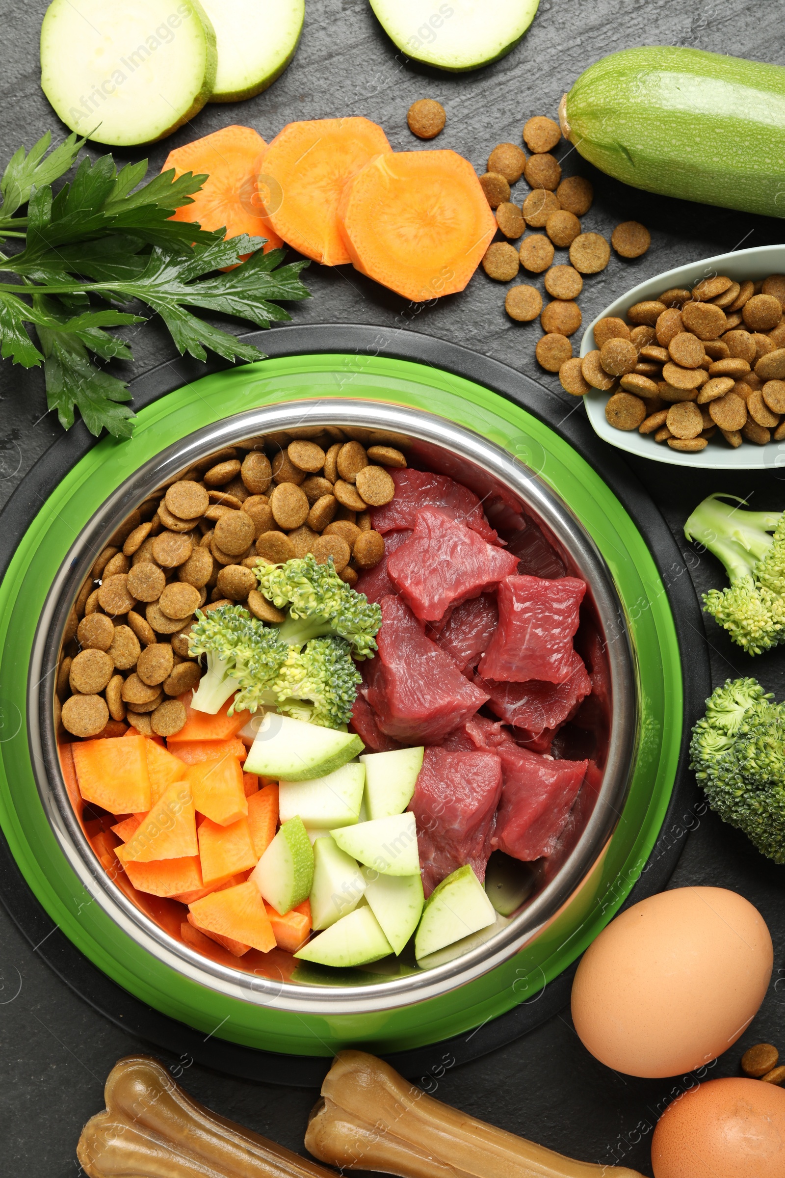 Photo of Natural and dry pet food in feeding bowl among products on dark textured table, flat lay