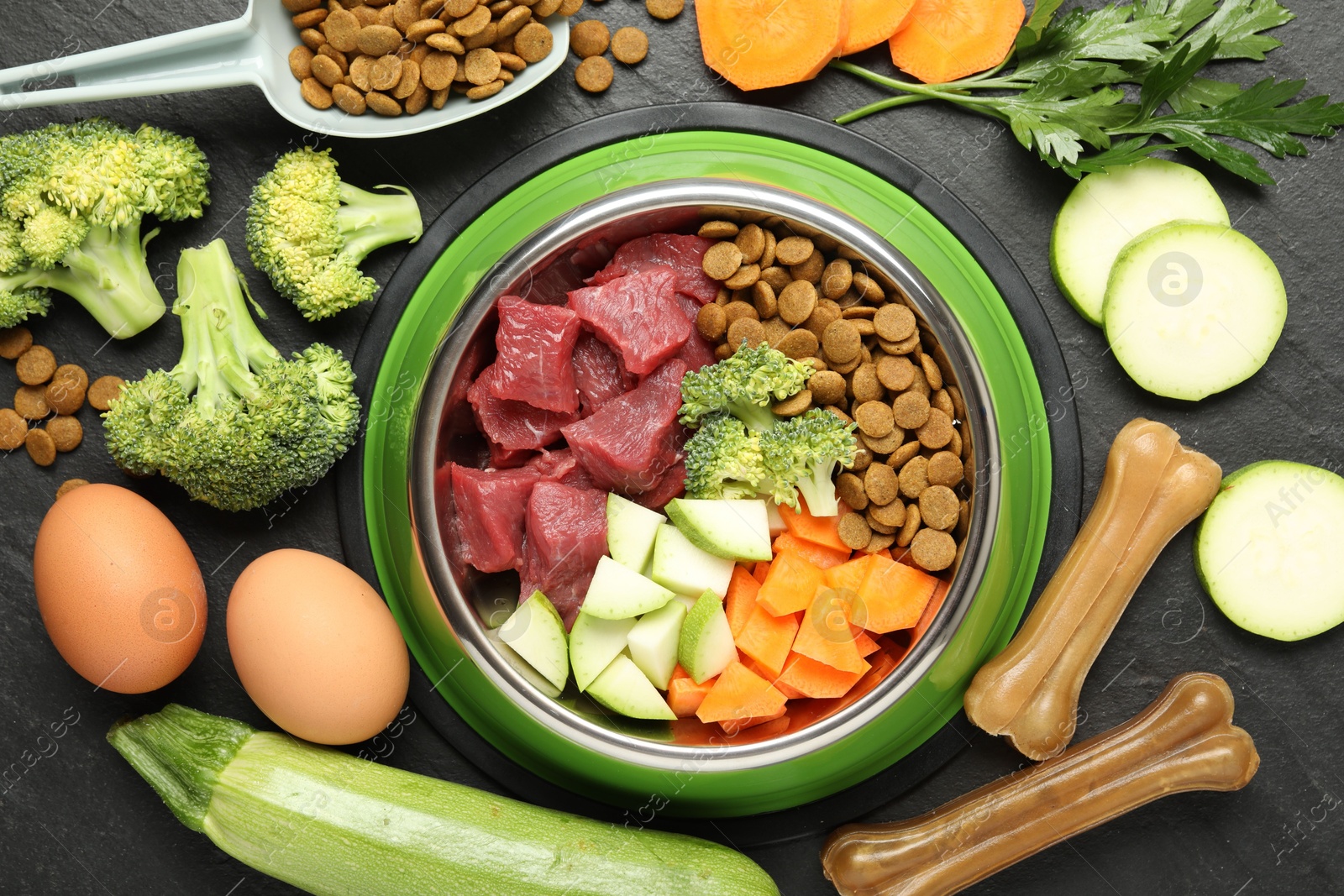 Photo of Natural and dry pet food in feeding bowl among products on dark textured table, flat lay