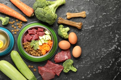 Photo of Natural and dry pet food in feeding bowls among products on dark textured table, flat lay. Space for text