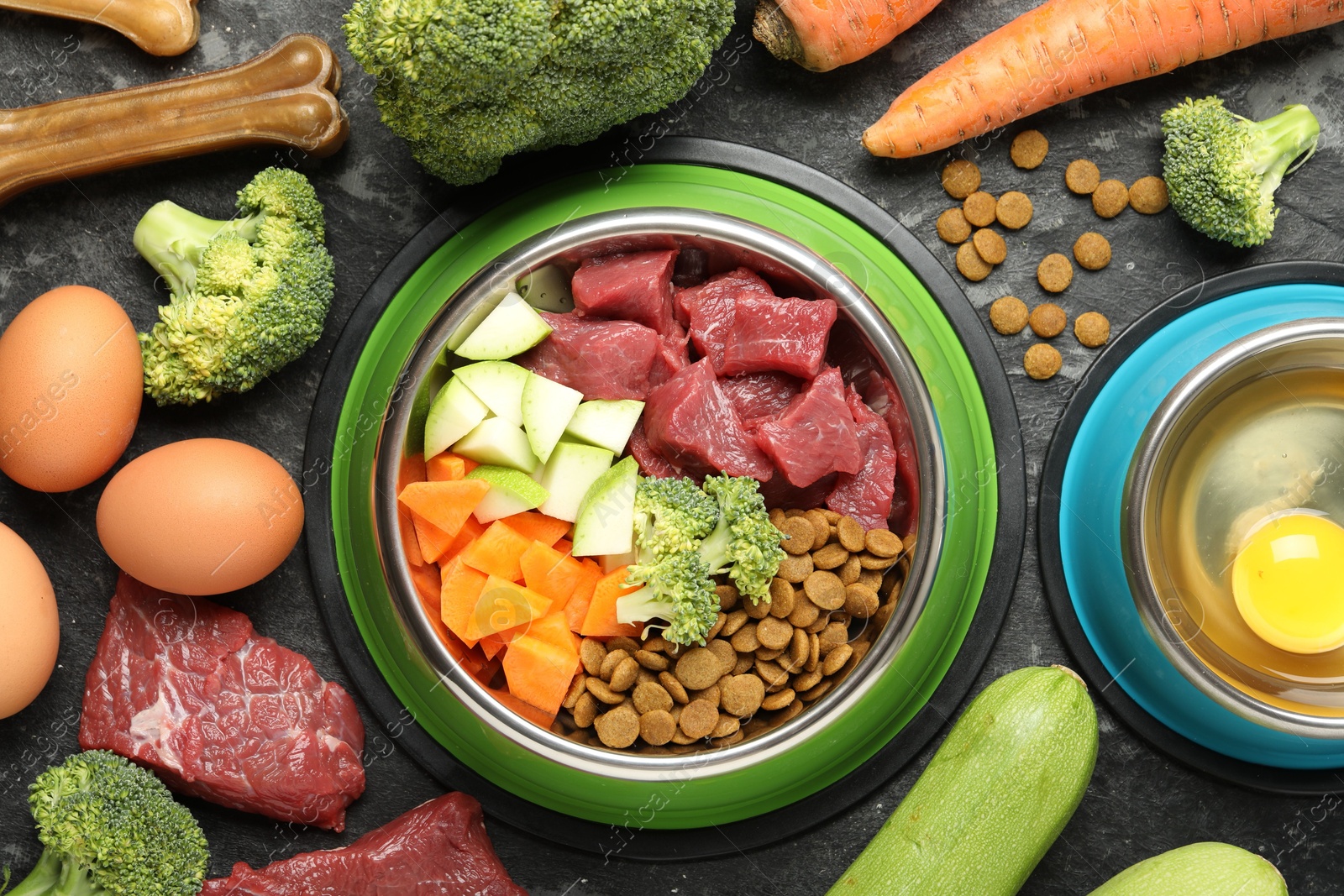 Photo of Natural and dry pet food in feeding bowls among products on dark textured table, flat lay