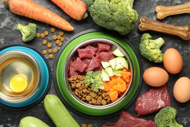 Photo of Natural and dry pet food in feeding bowls among products on dark textured table, flat lay