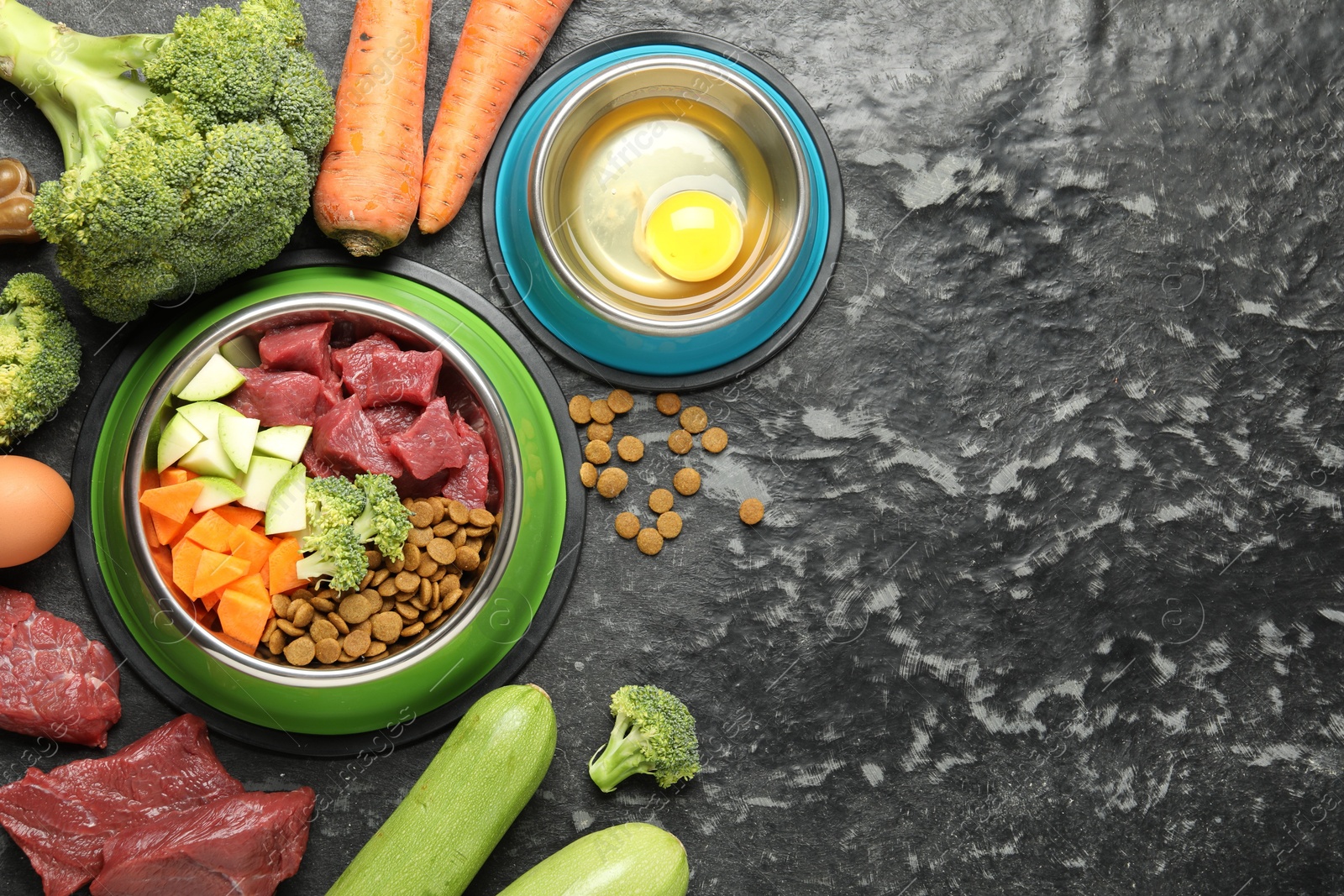 Photo of Natural and dry pet food in feeding bowls among products on dark textured table, flat lay. Space for text