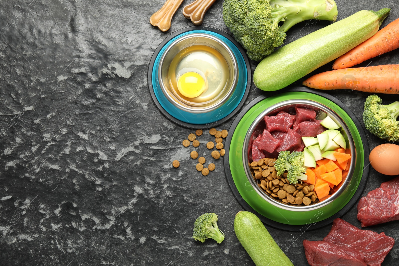 Photo of Natural and dry pet food in feeding bowls among products on dark textured table, flat lay. Space for text