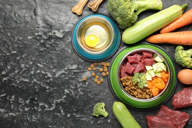 Photo of Natural and dry pet food in feeding bowls among products on dark textured table, flat lay. Space for text