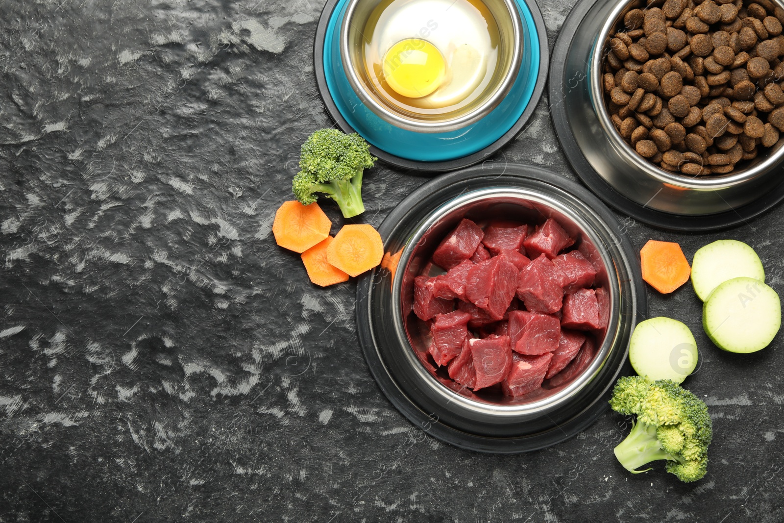 Photo of Natural and dry pet food in feeding bowls among products on dark textured table, flat lay. Space for text