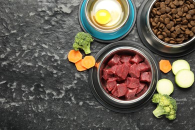 Photo of Natural and dry pet food in feeding bowls among products on dark textured table, flat lay. Space for text