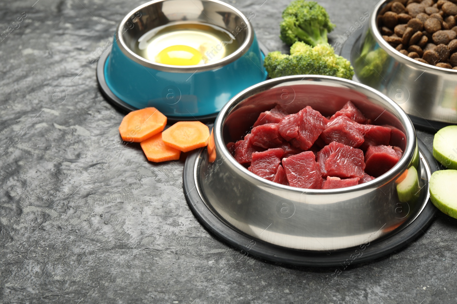 Photo of Natural and dry pet food in feeding bowls among products on dark textured table, closeup. Space for text