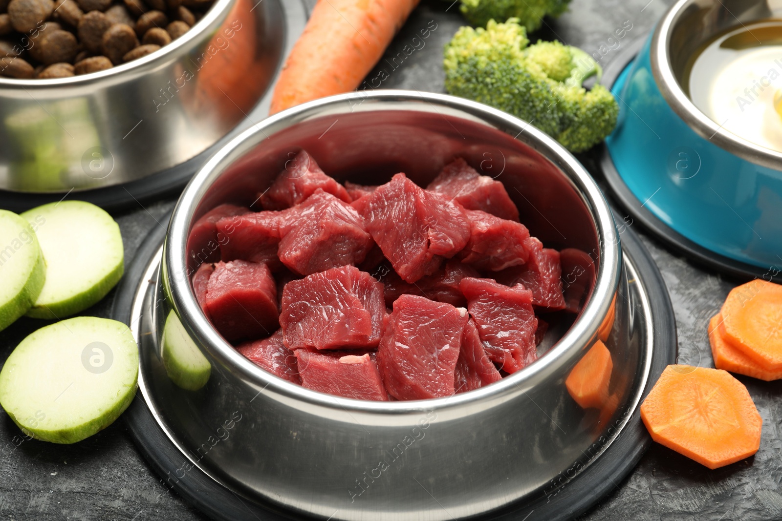 Photo of Natural and dry pet food in feeding bowls among products on dark textured table, closeup