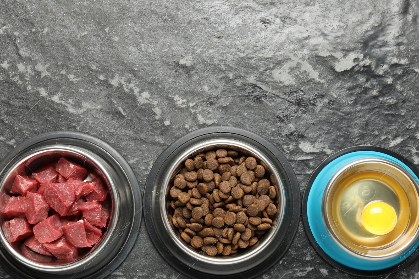 Photo of Natural and dry pet food in feeding bowls on dark textured table, flat lay. Space for text