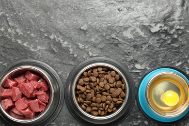 Photo of Natural and dry pet food in feeding bowls on dark textured table, flat lay. Space for text