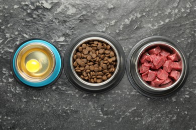 Photo of Natural and dry pet food in feeding bowls on dark textured table, flat lay