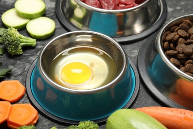 Photo of Natural and dry pet food in feeding bowls among products on dark textured table, closeup