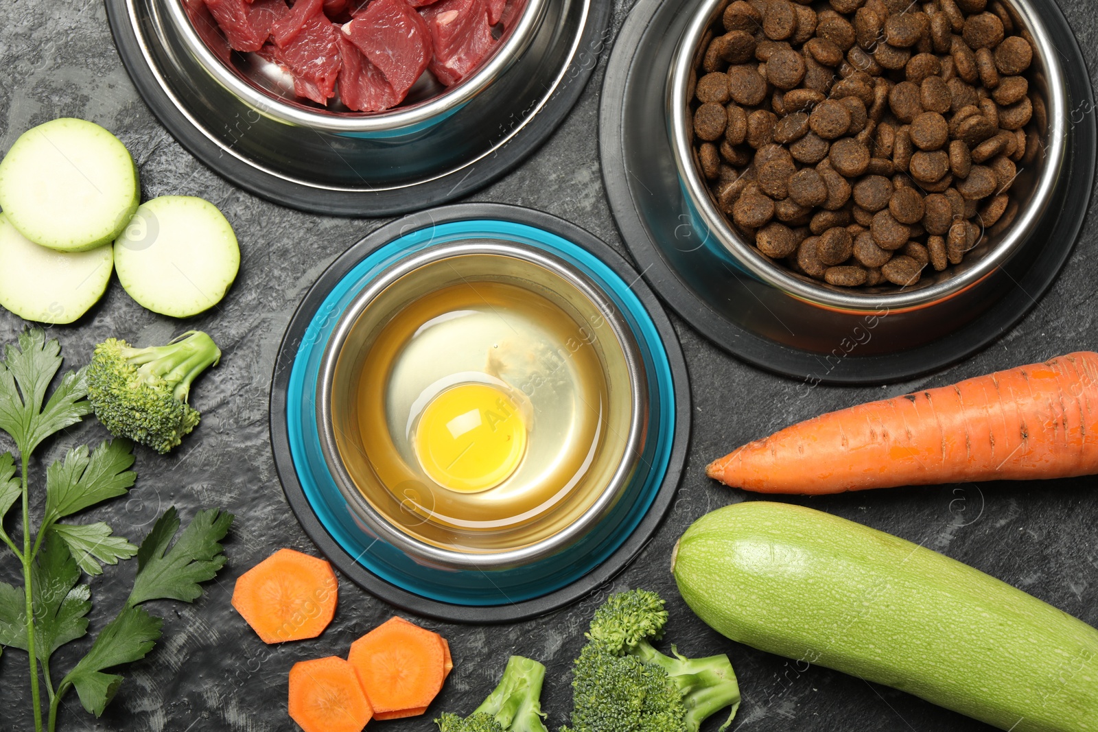 Photo of Natural and dry pet food in feeding bowls among products on dark textured table, flat lay