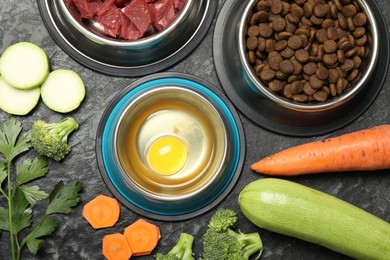 Photo of Natural and dry pet food in feeding bowls among products on dark textured table, flat lay