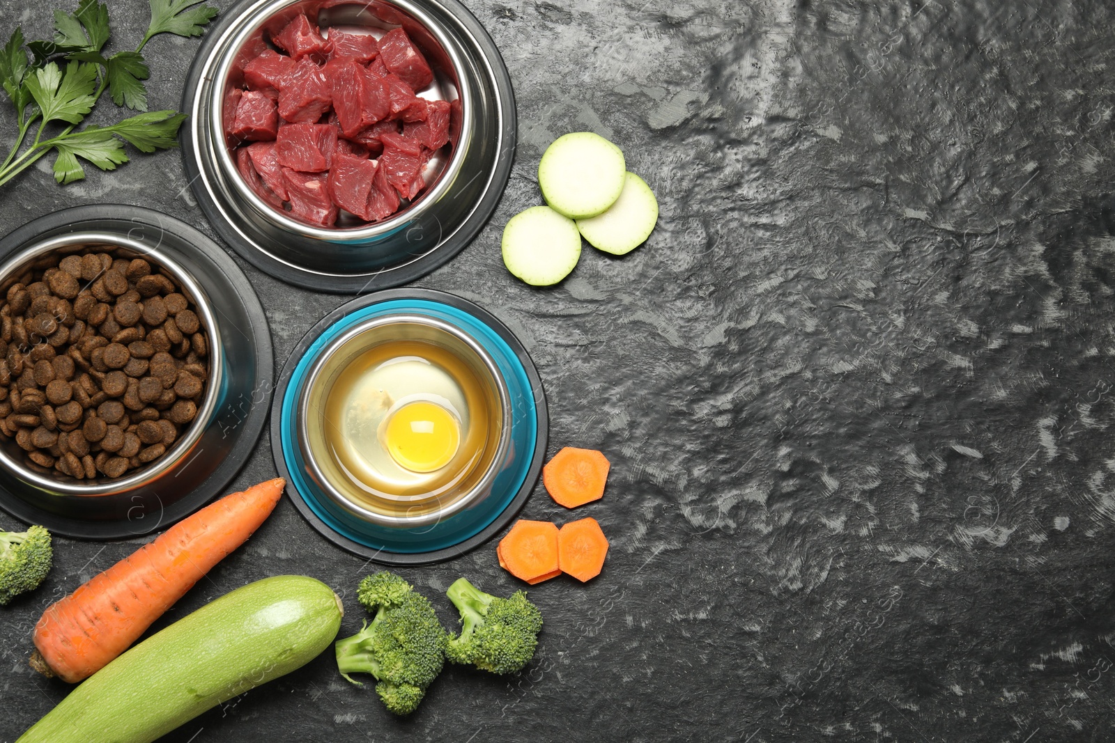 Photo of Natural and dry pet food in feeding bowls among products on dark textured table, flat lay. Space for text