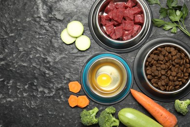 Photo of Natural and dry pet food in feeding bowls among products on dark textured table, flat lay. Space for text