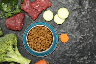 Photo of Dry pet food in feeding bowl and natural products on dark textured table, flat lay. Space for text