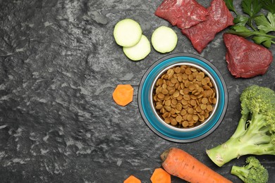 Photo of Dry pet food in feeding bowl and natural products on dark textured table, flat lay. Space for text