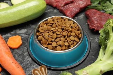 Photo of Dry pet food in feeding bowl and natural products on dark textured table, closeup