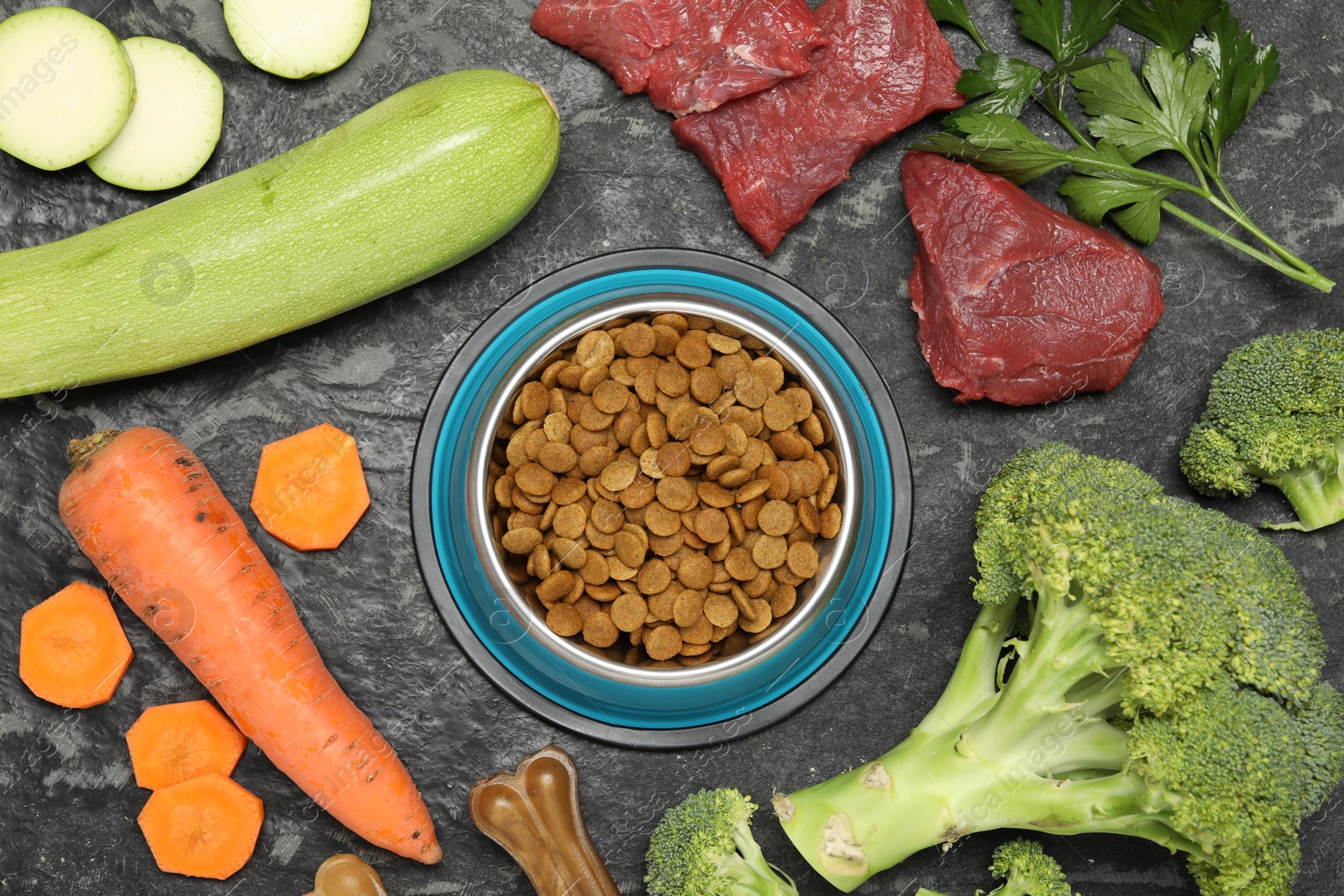 Photo of Dry pet food in feeding bowl and natural products on dark textured table, flat lay