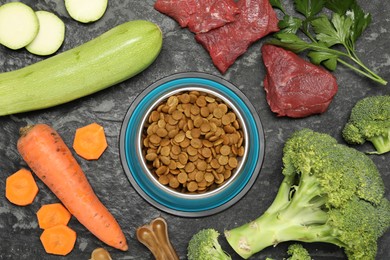 Photo of Dry pet food in feeding bowl and natural products on dark textured table, flat lay