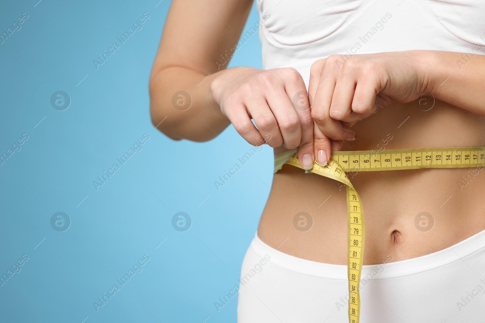 Photo of Weight loss. Woman measuring her slim waist with tape on light blue background, closeup. Space for text