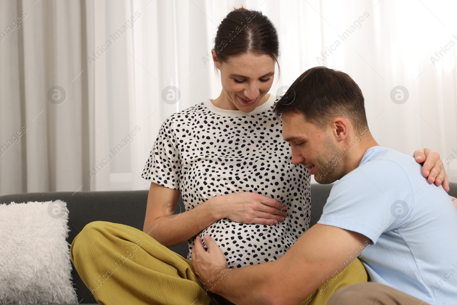 Photo of Man trying to feel baby kicks in his pregnant wife's belly on sofa at home