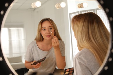 Photo of Beautiful young woman applying eye shadow with brush near mirror indoors, view through ring lamp