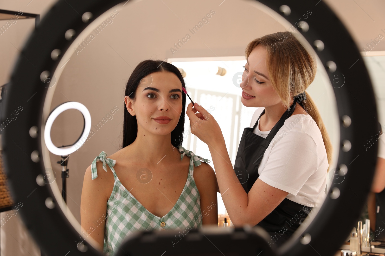 Photo of Professional makeup artist working with beautiful young woman in salon, view through ring lamp