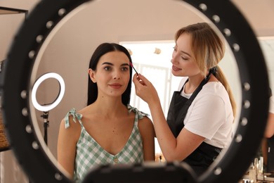 Photo of Professional makeup artist working with beautiful young woman in salon, view through ring lamp