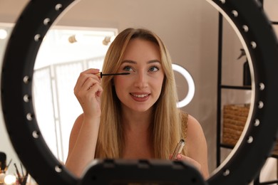 Photo of Beautiful young woman applying mascara indoors, view through ring lamp