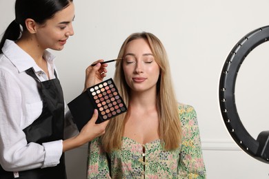 Photo of Professional makeup artist working with beautiful young woman near white wall. Using ring lamp