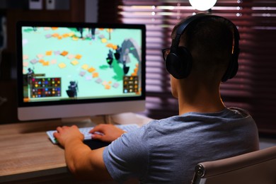 Photo of Young man playing video game with keyboard at wooden table indoors, back view