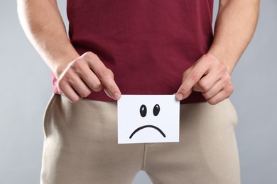 Photo of STD (sexually transmitted diseases). Man holding sticky note with drawn sad face on grey background, closeup