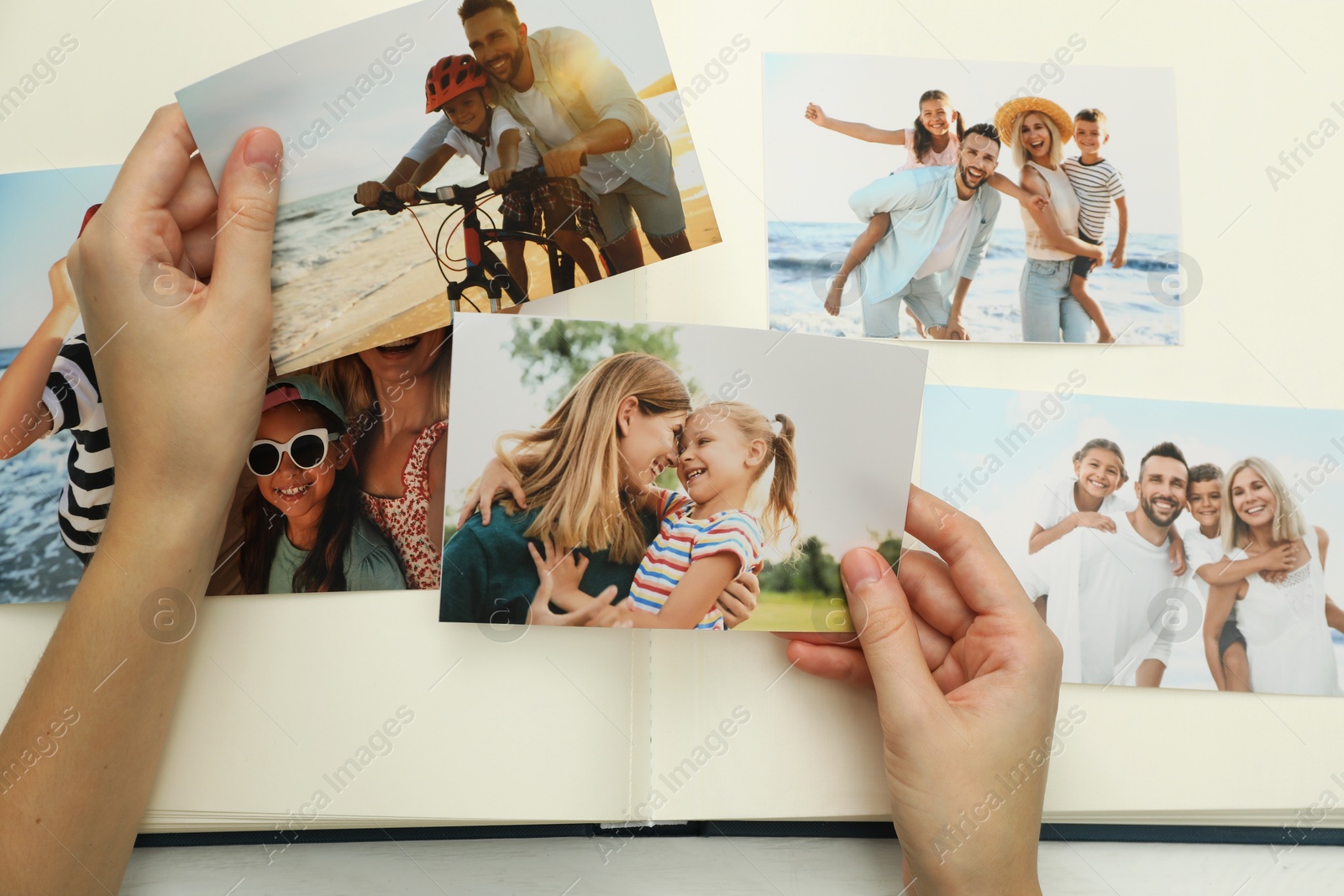 Photo of Woman with different photos in open photo album at white wooden table, top view