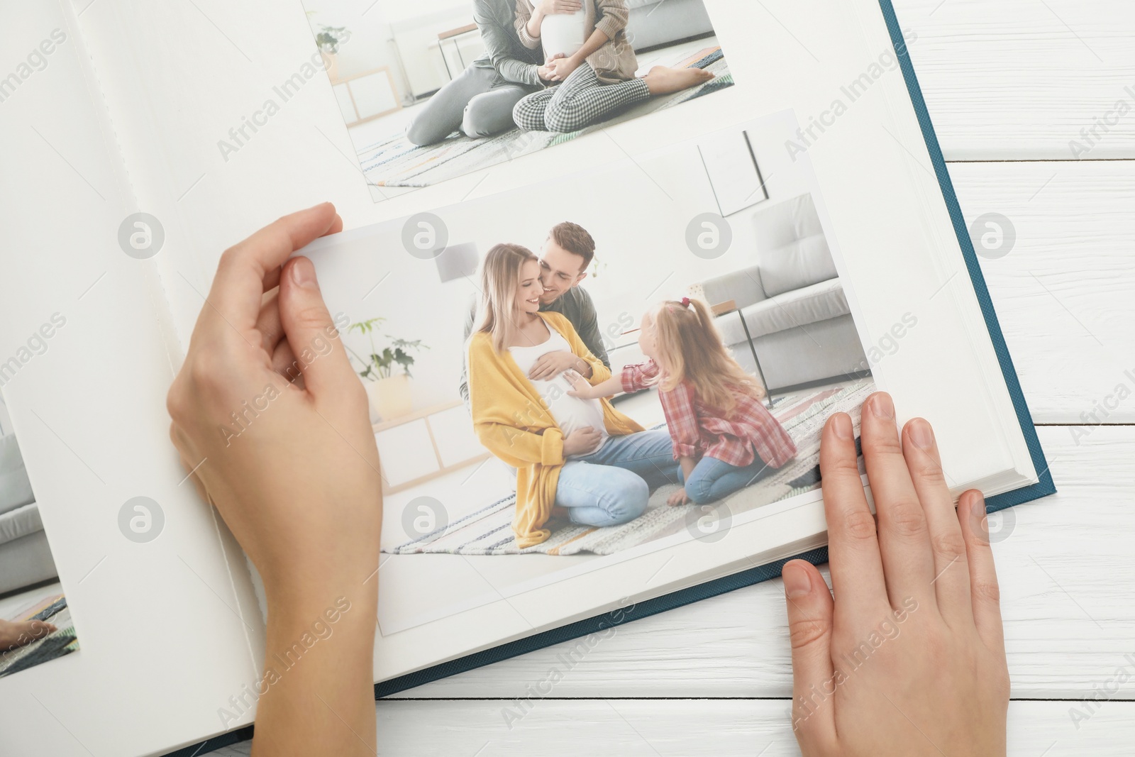 Photo of Woman with different photos in open photo album at white wooden table, top view