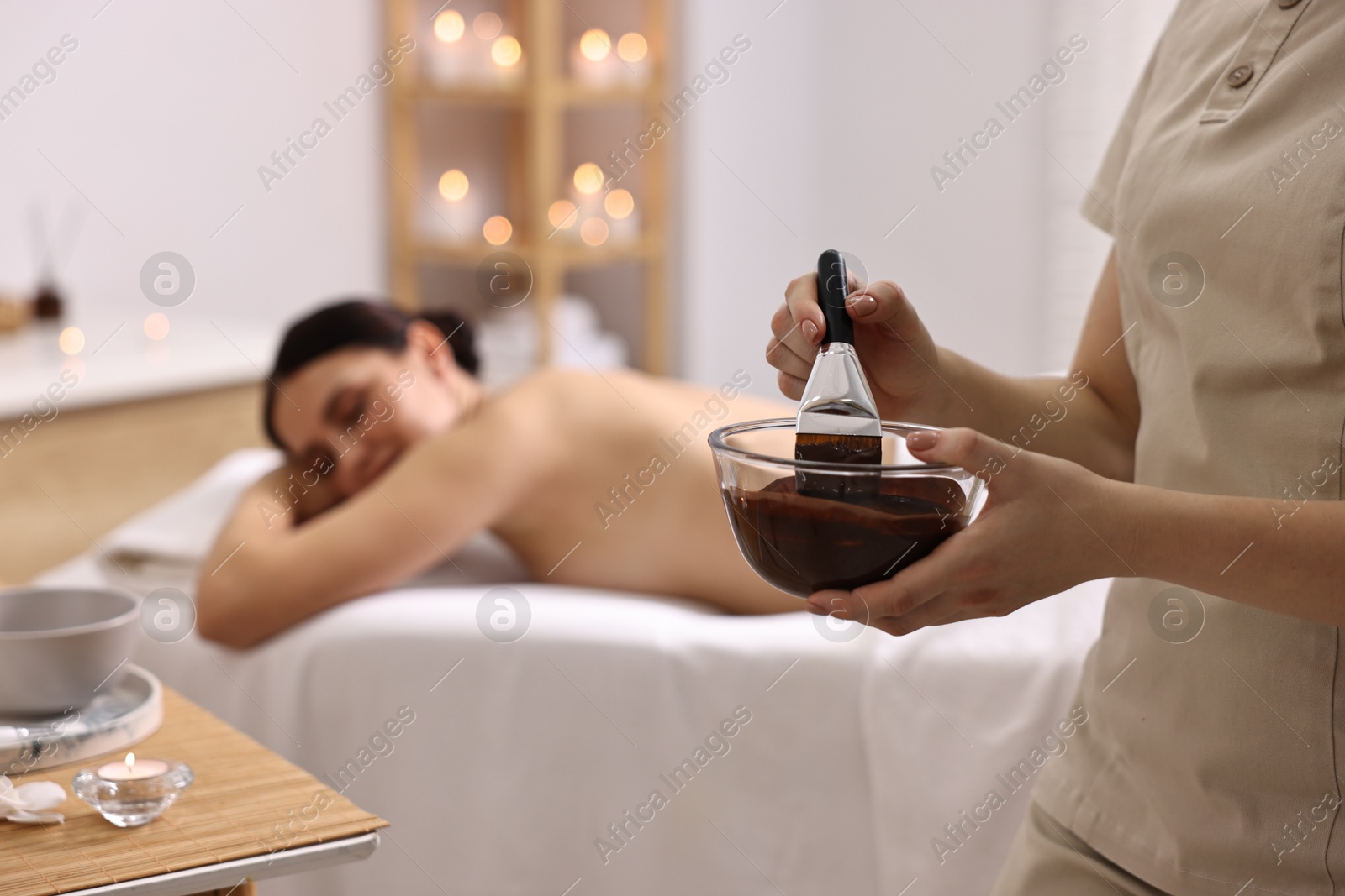 Photo of Spa worker with cosmetic product for chocolate body wrap near woman in salon, closeup. Selective focus