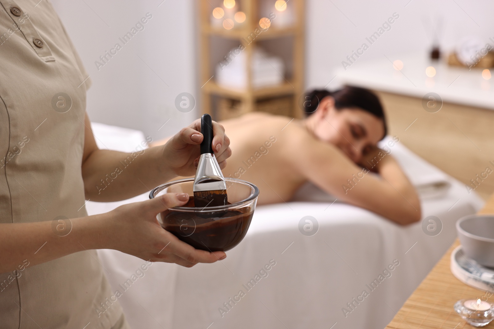 Photo of Spa worker with cosmetic product for chocolate body wrap near woman in salon, closeup. Selective focus