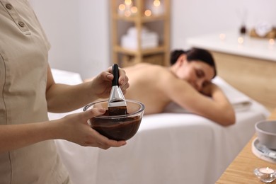 Photo of Spa worker with cosmetic product for chocolate body wrap near woman in salon, closeup. Selective focus