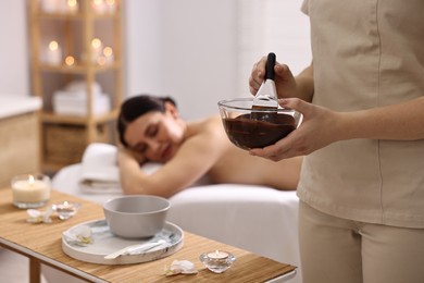 Photo of Spa worker with cosmetic product for chocolate body wrap near woman in salon, closeup. Selective focus