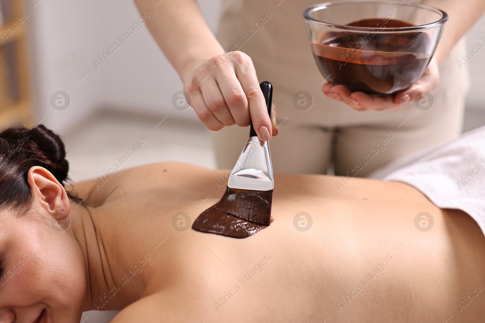 Photo of Chocolate body wrap. Spa worker applying mask onto woman's back in salon, closeup