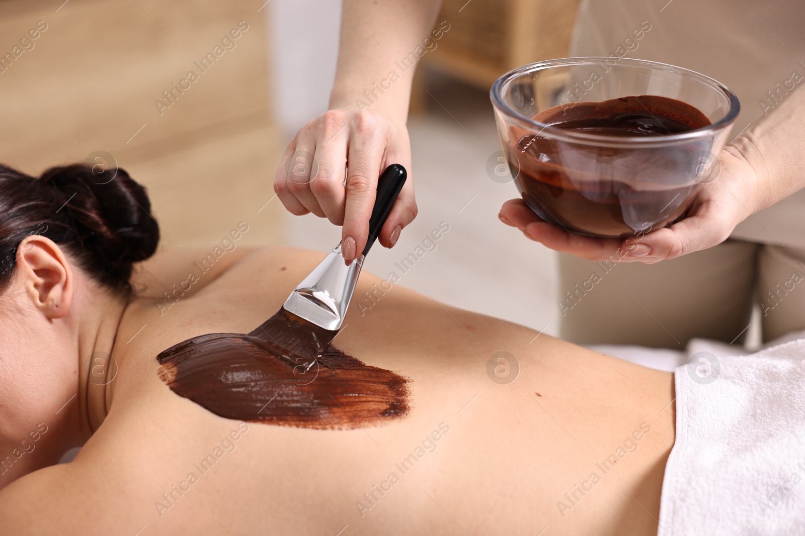 Photo of Chocolate body wrap. Spa worker applying mask onto woman's back in salon, closeup