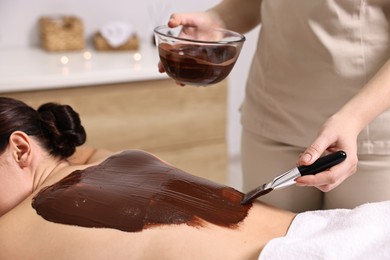 Photo of Chocolate body wrap. Spa worker applying mask onto woman's back in salon, closeup