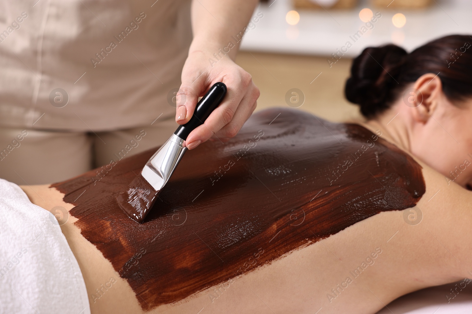 Photo of Chocolate body wrap. Spa worker applying mask onto woman's back in salon, closeup