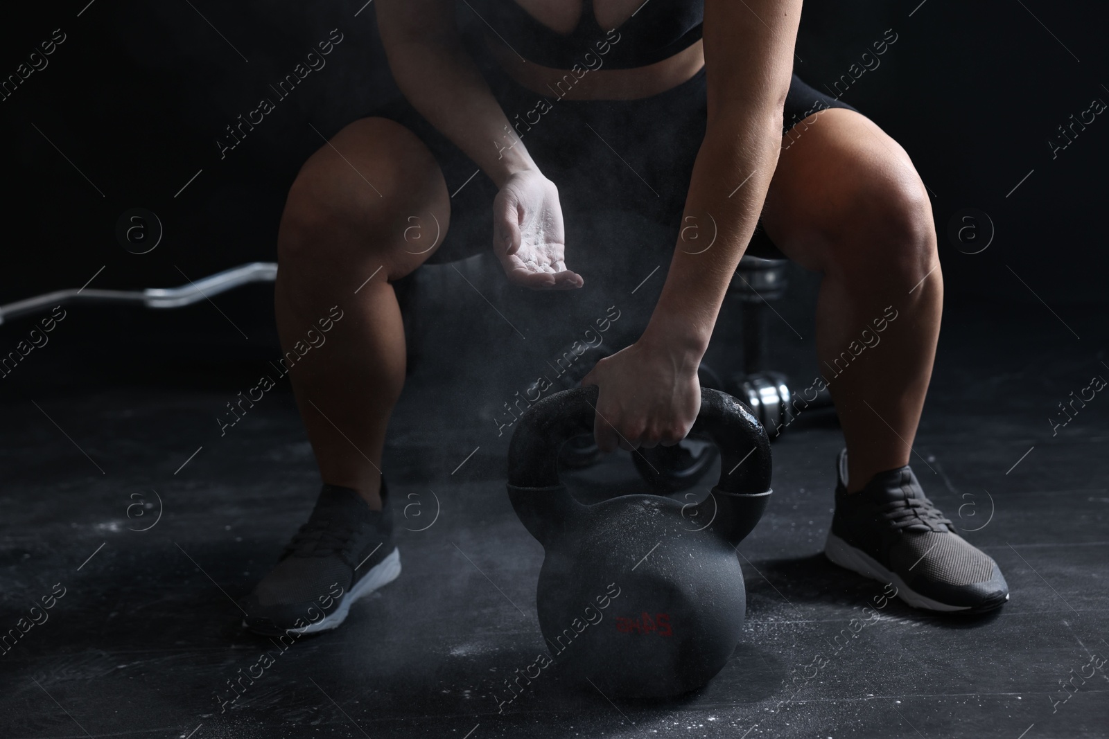 Photo of Woman with talcum powder on hands training with kettlebell against black background, closeup