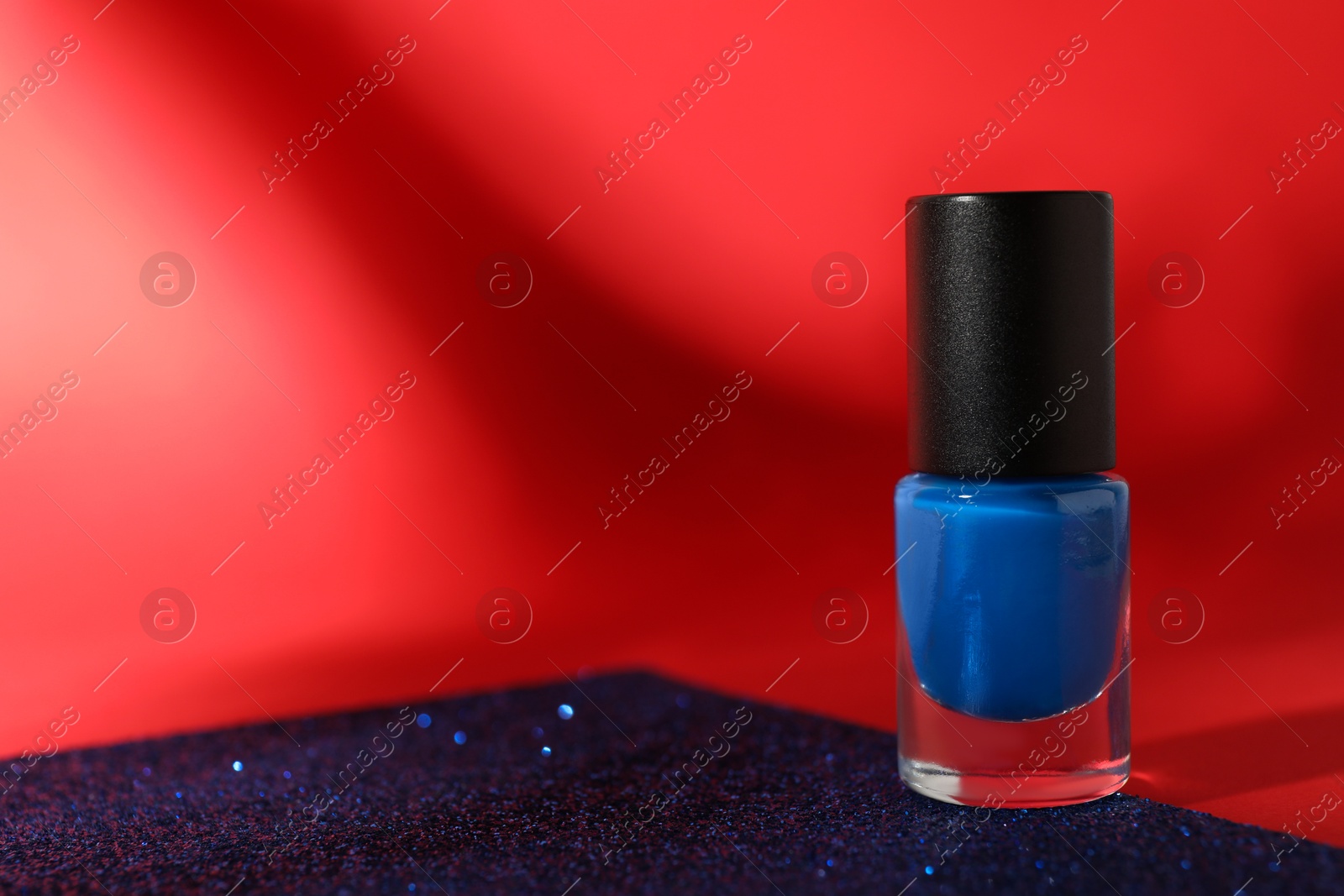 Photo of Blue nail polish in bottle on bright glitter surface against red background, space for text