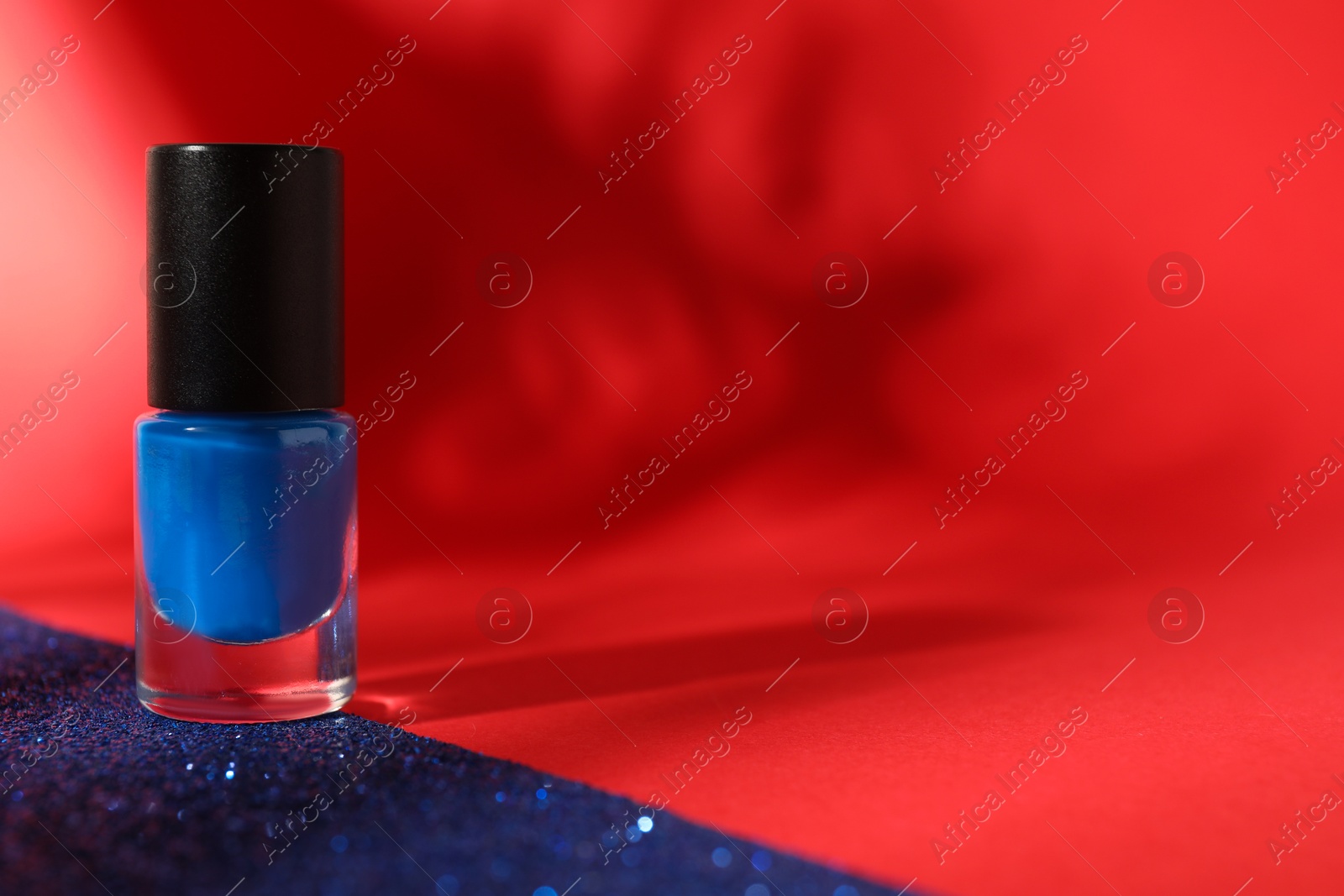 Photo of Blue nail polish in bottle on bright glitter surface against red background, space for text