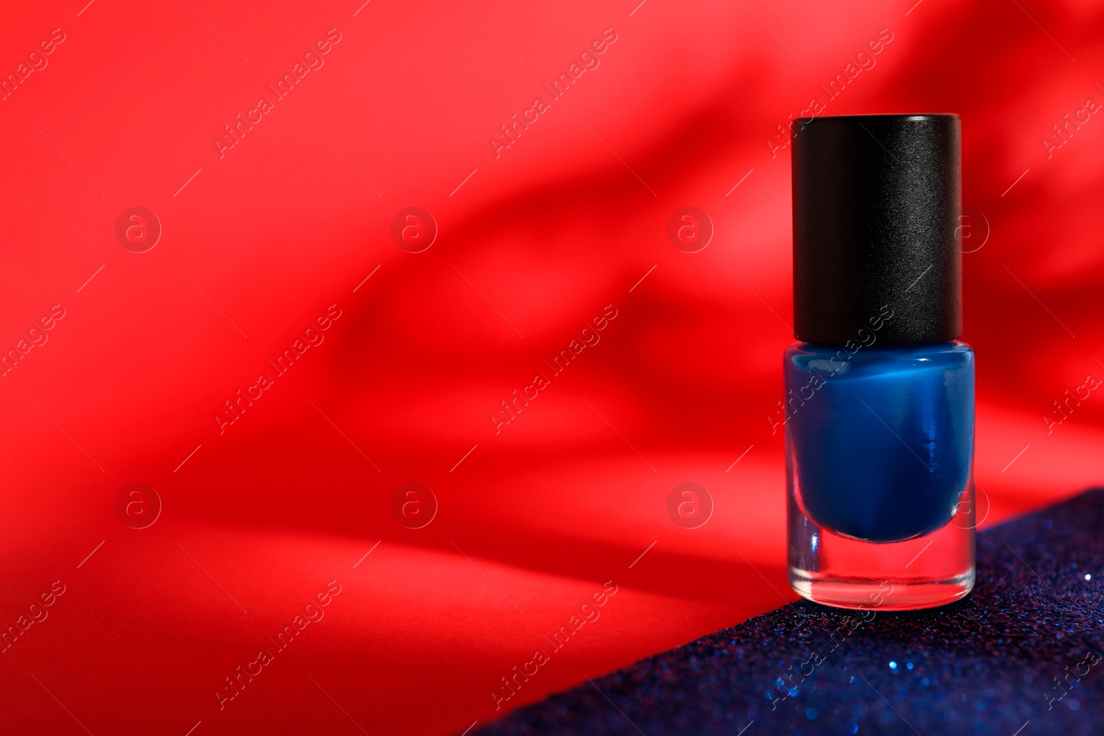 Photo of Blue nail polish in bottle on bright glitter surface against red background, space for text