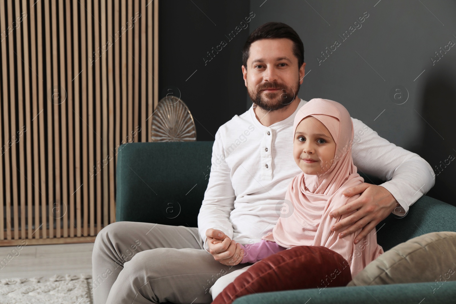 Photo of Muslim man and his daughter sitting on sofa at home, space for text